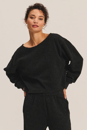 A person with short curly hair wears the MALLORY SWEATSHIRT by Velvet by Graham & Spencer and matching black pants, standing against a plain light background.