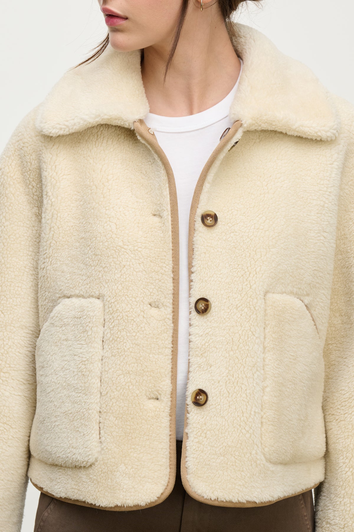   A woman wearing the AMARA SHERPA REVERSIBLE JACKET by Velvet by Graham & Spencer—a cream faux sherpa jacket with large front pockets and brown buttons—over a white shirt and brown pants. 