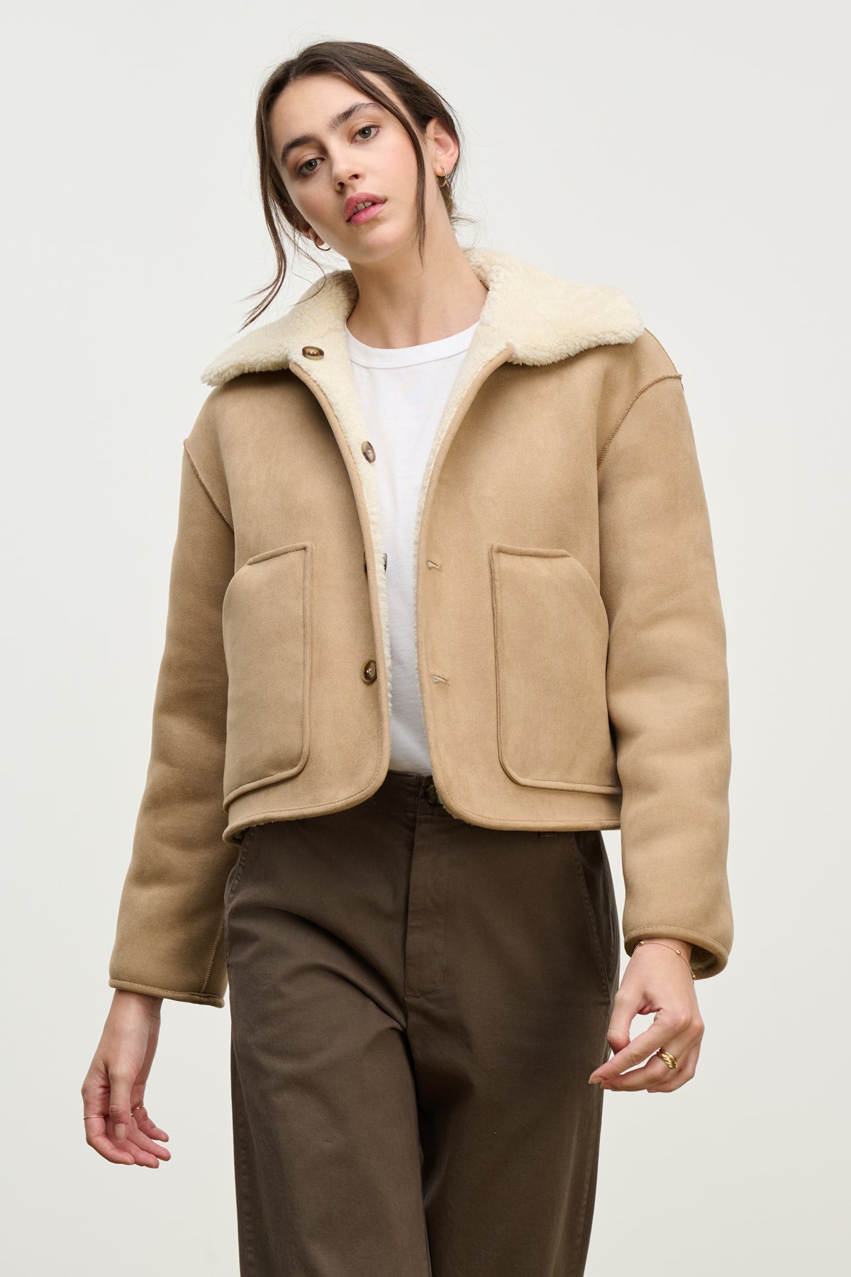   A woman wears the AMARA SHERPA REVERSIBLE JACKET by Velvet by Graham & Spencer over a white shirt and brown pants, standing against a plain light background. 