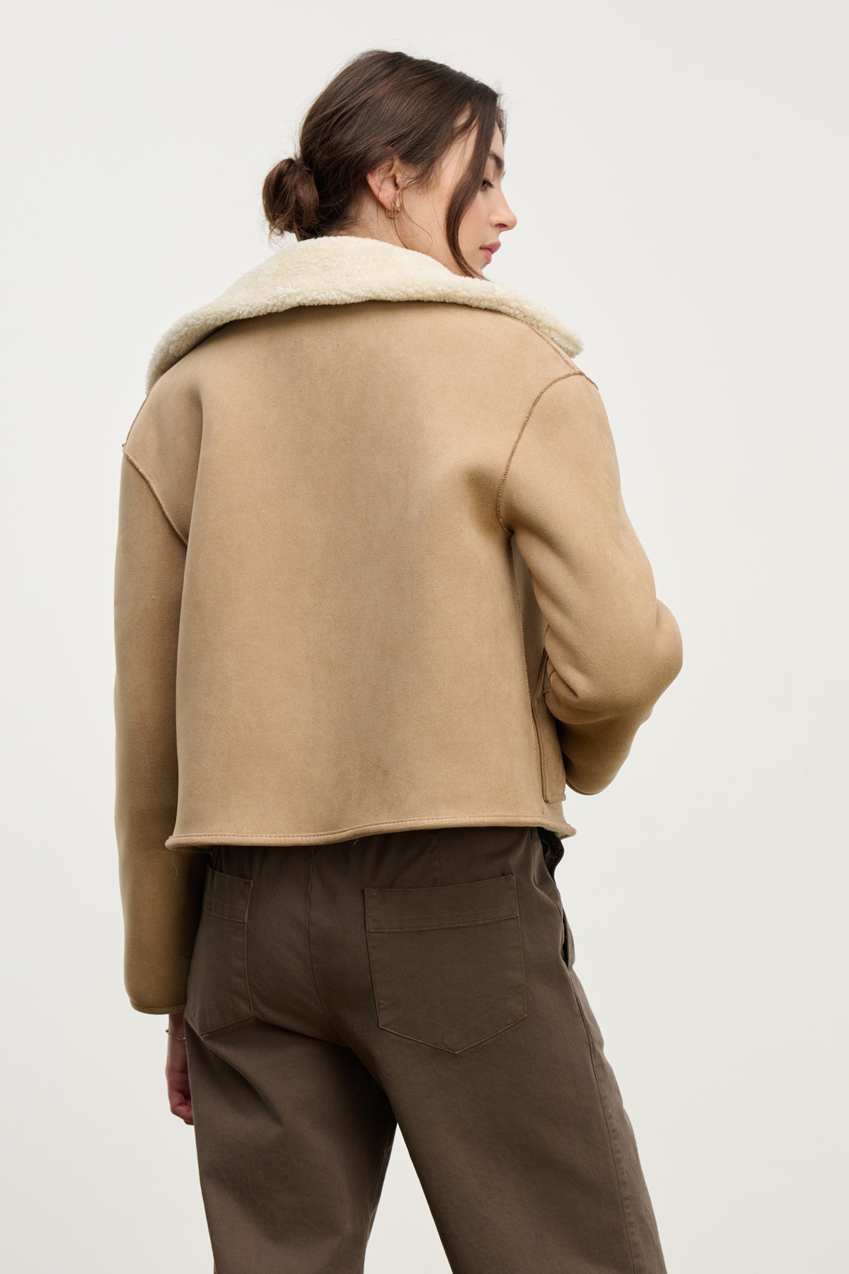   A woman with brown hair in a low bun faces away from the camera, wearing Velvet by Graham & Spencer’s AMARA SHERPA REVERSIBLE JACKET and brown pants against a plain light background. 