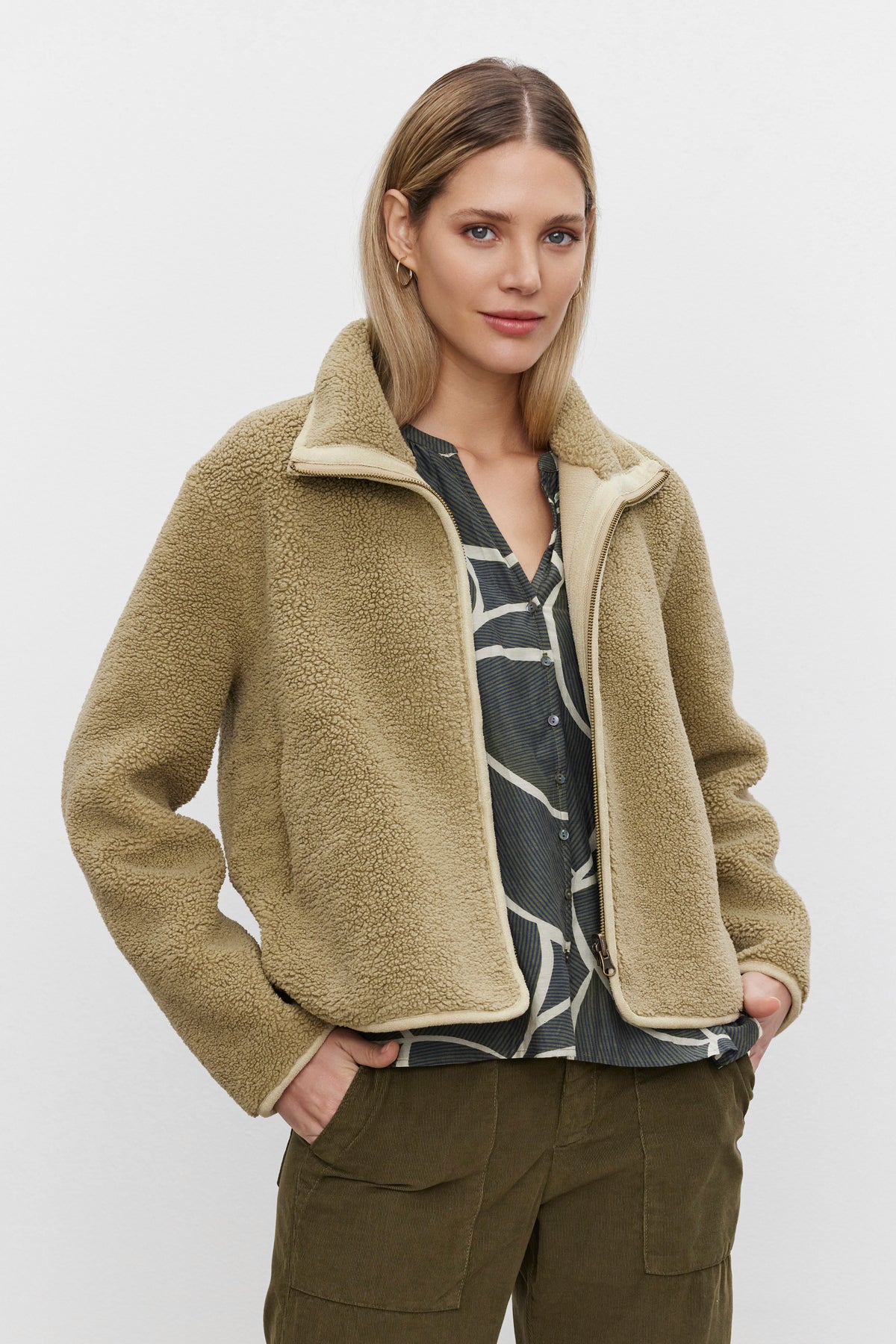   A person with blonde hair wears the REBA REVERSIBLE SHERPA JACKET by Velvet by Graham & Spencer over a patterned shirt and brown pants, standing against a plain white background. 