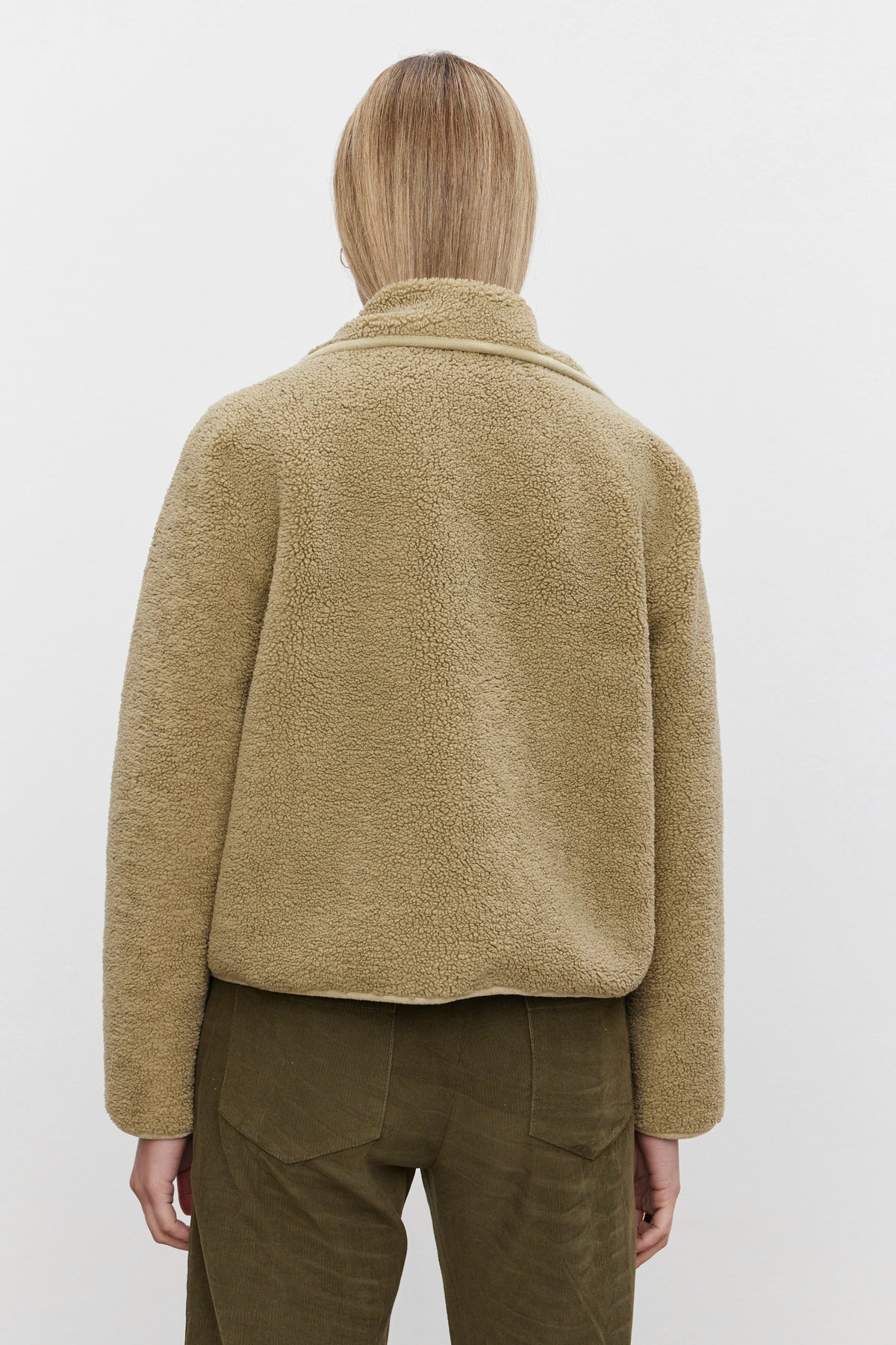   Person with shoulder-length blonde hair wearing a beige REBA REVERSIBLE SHERPA JACKET by Velvet by Graham & Spencer and green pants, seen from behind. 