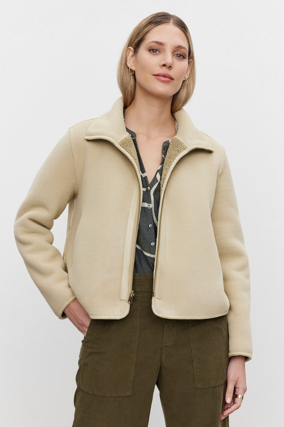   A person with shoulder-length hair wears the cozy REBA REVERSIBLE SHERPA JACKET by Velvet by Graham & Spencer over a grey shirt and green pants, standing against a plain white background. 