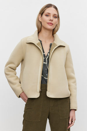 A person with shoulder-length hair wears the cozy REBA REVERSIBLE SHERPA JACKET by Velvet by Graham & Spencer over a grey shirt and green pants, standing against a plain white background.