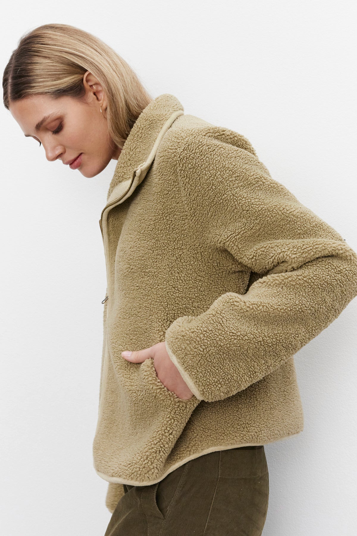   A person with short, blonde hair wears the REBA REVERSIBLE SHERPA JACKET by Velvet by Graham & Spencer in beige and has their left hand in their pocket. They are looking down against a plain white background, enjoying the cozy feel. 