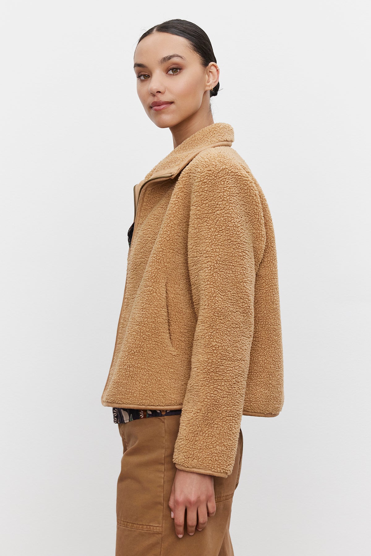   A woman wearing the REBA REVERSIBLE SHERPA JACKET by Velvet by Graham & Spencer, featuring a tan faux sherpa with faux suede trims, stands against a plain background, looking over her shoulder. She has her hair pulled back and is dressed in brown pants. 