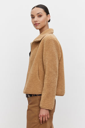 A woman wearing the REBA REVERSIBLE SHERPA JACKET by Velvet by Graham & Spencer, featuring a tan faux sherpa with faux suede trims, stands against a plain background, looking over her shoulder. She has her hair pulled back and is dressed in brown pants.