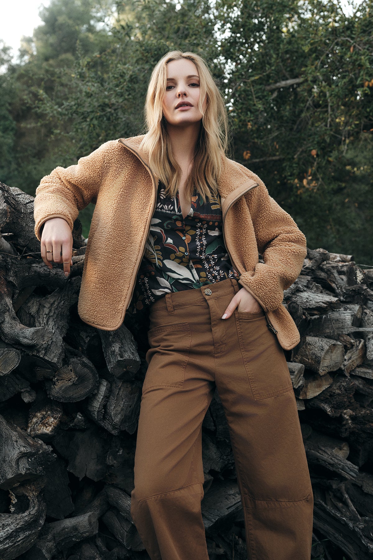   A person stands outdoors in front of a pile of logs, wearing the REBA REVERSIBLE SHERPA JACKET by Velvet by Graham & Spencer over a floral shirt and brown pants. Trees are visible in the background. 