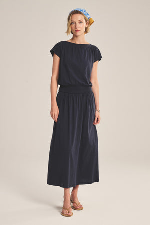 A woman wears the Velvet by Graham & Spencer SHILOH SKIRT in navy blue with a short-sleeve top, tan sandals, and a blue patterned headband, set against a plain beige background.