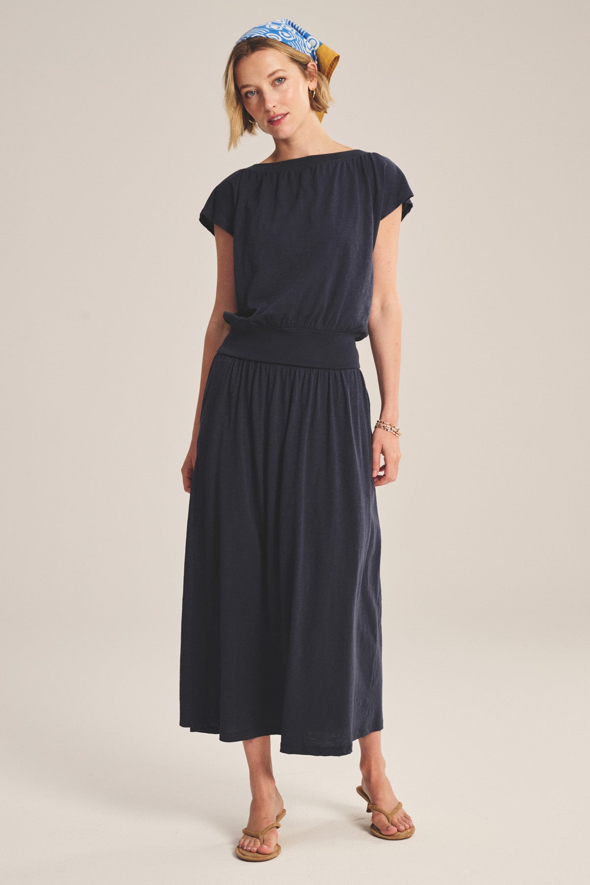 A woman wears the SHILOH SKIRT by Velvet by Graham & Spencer, a navy A-line midi with fitted waistband, styled with tan sandals and a blue patterned headscarf against a neutral background.-41296999219393