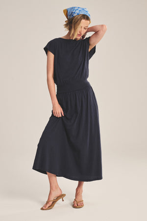 A woman wearing the SHILOH SKIRT by Velvet by Graham & Spencer, tan sandals, and a blue patterned headscarf stands with one hand touching her head.