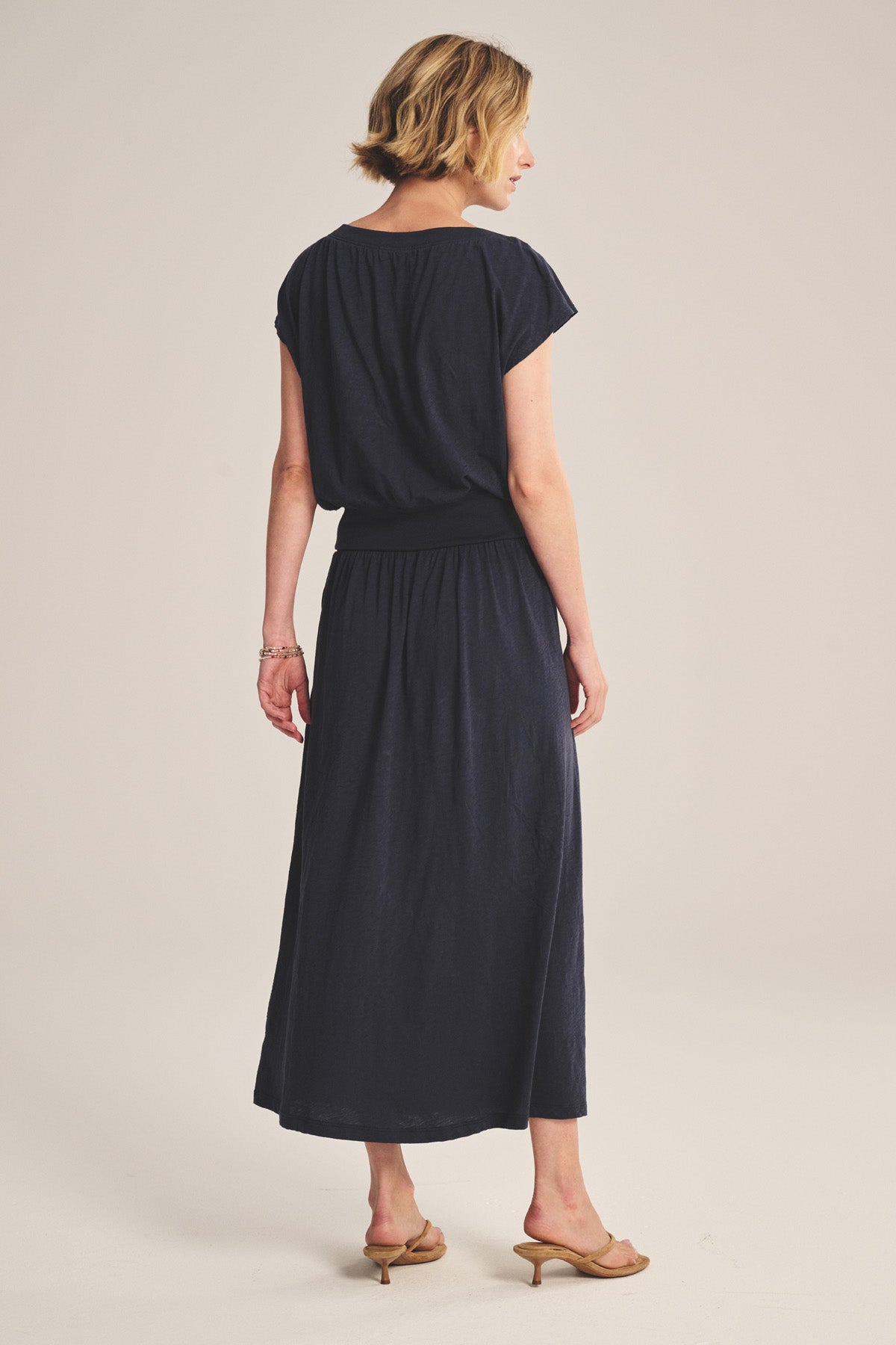 A woman faces away, wearing the SHILOH SKIRT by Velvet by Graham & Spencer—navy, A-line, midi to maxi length—styled as a short-sleeve ankle-length dress with beige heeled sandals against a light background.-41296999088321