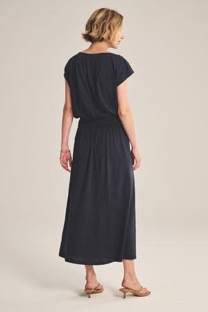 A woman faces away, wearing the SHILOH SKIRT by Velvet by Graham & Spencer—navy, A-line, midi to maxi length—styled as a short-sleeve ankle-length dress with beige heeled sandals against a light background.