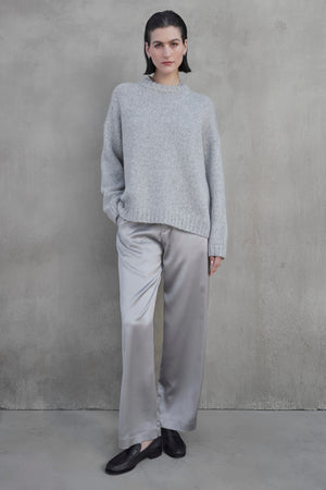 A person stands against a gray wall, wearing a gray CAMBRIA ALPACA SWEATER from Velvet by Jenny Graham, silver satin pants, and black loafers. Their left hand is in their pocket.