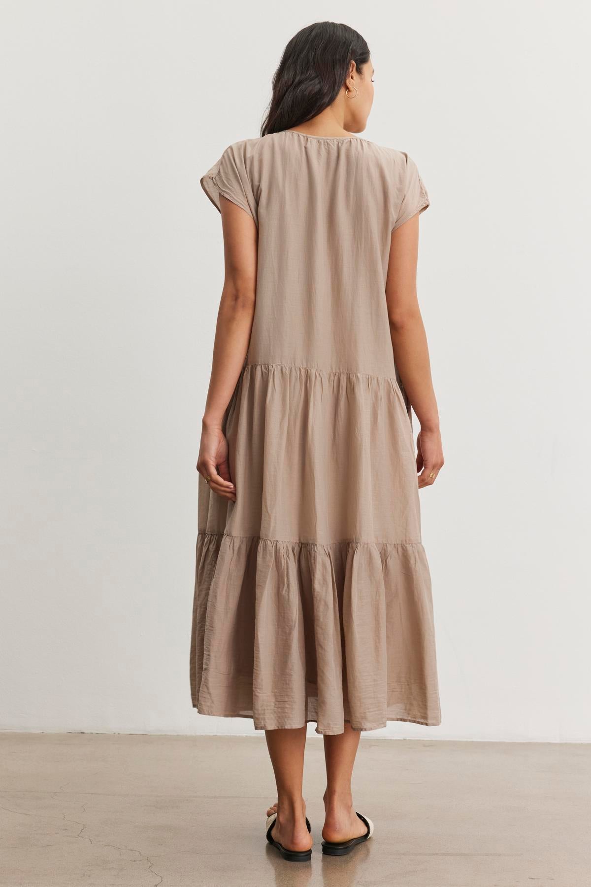   A woman stands with her back facing the camera, wearing the ADA SILK COTTON VOILE DRESS by Velvet by Graham & Spencer, which is a beige, tiered, knee-length dress made of silk cotton voile. She pairs it with sandals and is positioned against a plain, light-colored background. 