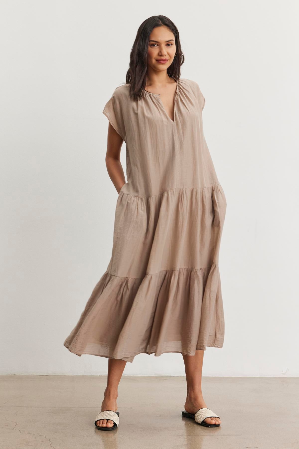   A woman stands wearing the Velvet by Graham & Spencer ADA Silk Cotton Voile Dress in light brown. The ankle-length dress features a relaxed silhouette and she pairs it with white sandals. With her hands tucked into the dress pockets, she smiles slightly while posing against a plain white background. 