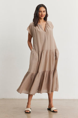 A woman stands wearing the Velvet by Graham & Spencer ADA Silk Cotton Voile Dress in light brown. The ankle-length dress features a relaxed silhouette and she pairs it with white sandals. With her hands tucked into the dress pockets, she smiles slightly while posing against a plain white background.