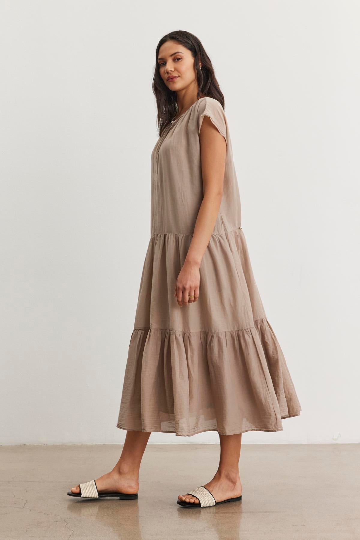 A woman stands against a plain background, elegantly showcasing the relaxed silhouette of the ADA SILK COTTON VOILE DRESS in light brown by Velvet by Graham & Spencer, paired gracefully with white sliders.-37054598447297