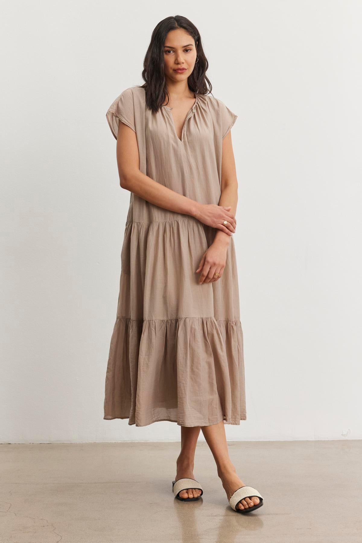   A person stands indoors wearing the ADA SILK COTTON VOILE DRESS in beige by Velvet by Graham & Spencer, paired with white slide sandals, with a neutral expression on their face. 