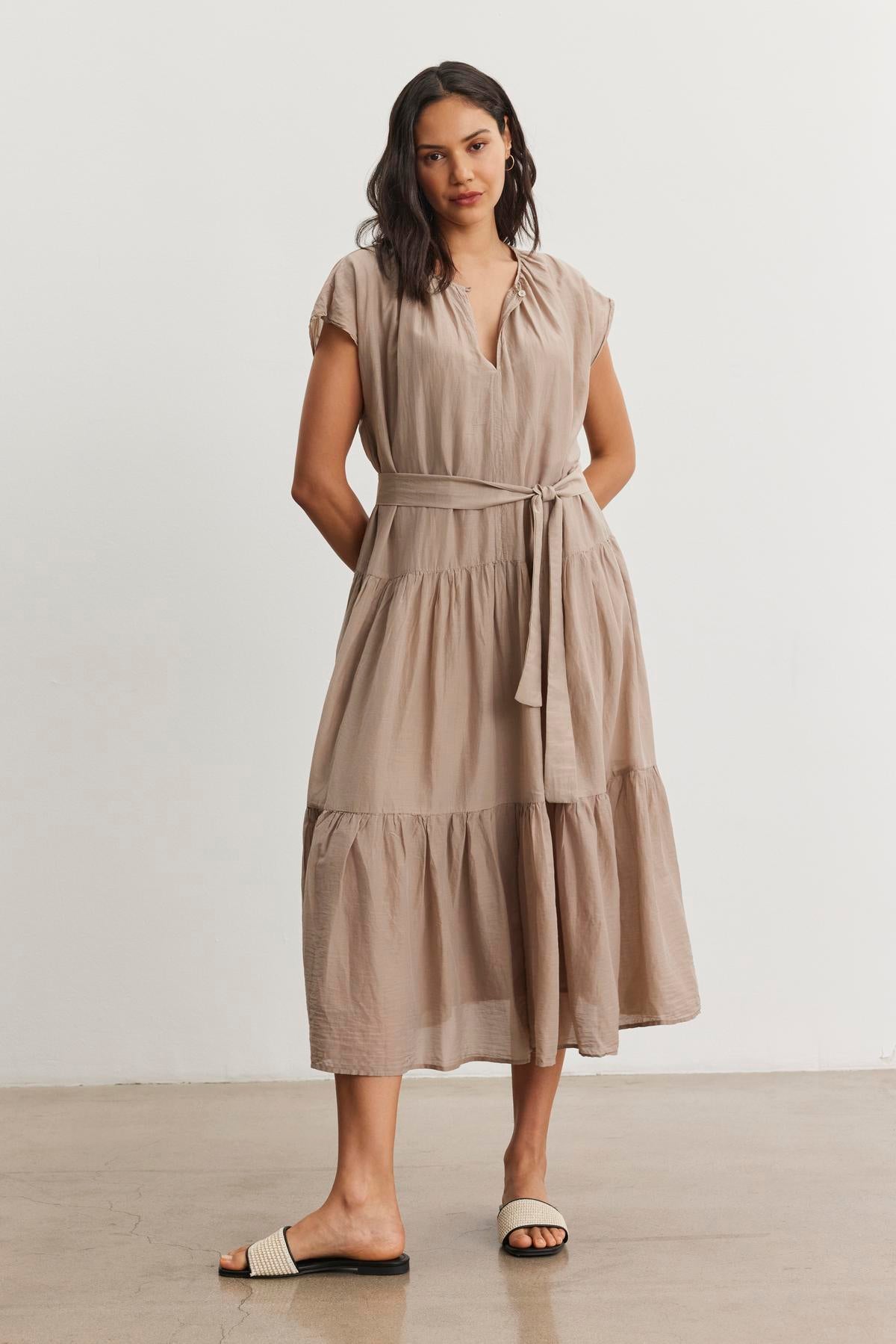   A woman stands against a plain white background, wearing the ADA SILK COTTON VOILE DRESS by Velvet by Graham & Spencer. The beige, short-sleeved, tiered dress features a tie at the waist and is paired with black and beige sandals. The relaxed silhouette of her outfit complements her shoulder-length dark hair perfectly. 