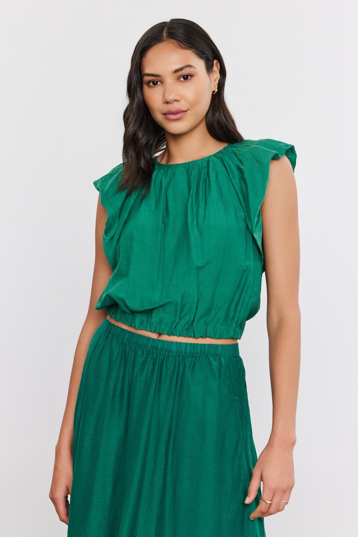 A woman in a green ruffled sleeveless AMORA TOP with a crew neckline and matching skirt, standing against a white background. (Brand: Velvet by Graham & Spencer)-36910240366785