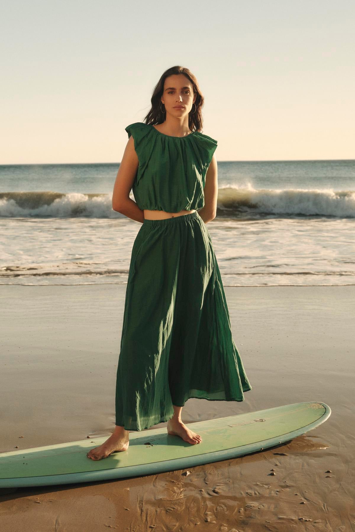 A woman in a green silk dress stands on a surfboard on the beach, with the ocean in the background, wearing an AMORA TOP from Velvet by Graham & Spencer.-36910240530625