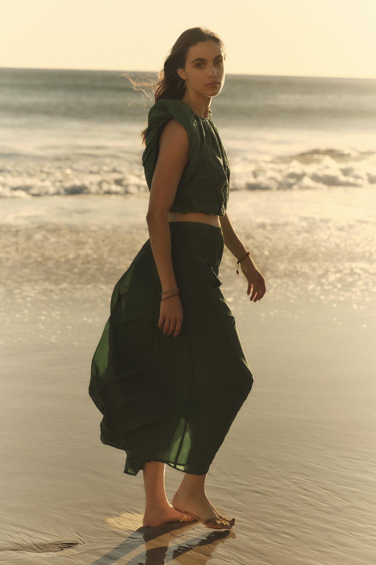   A woman in a flowing green Amora Top by Velvet by Graham & Spencer walks barefoot along a beach at sunset, looking over her shoulder. 