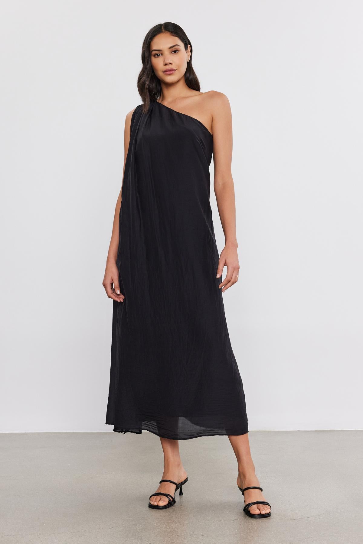 Woman in a black one-shoulder Velvet by Graham & Spencer DIANA DRESS and black heels, standing on a plain backdrop.-36918818898113
