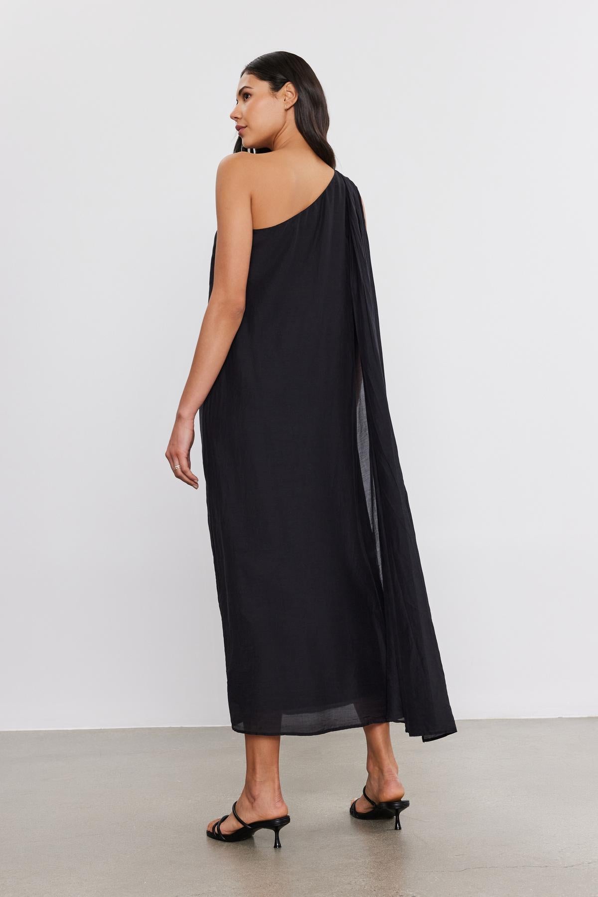 A woman stands with her back to the camera, wearing a flowing black one-shoulder Diana Dress made of silk cotton voile by Velvet by Graham & Spencer and black high heels.-36918818963649