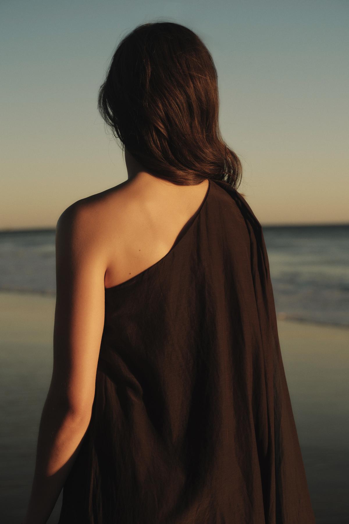   A woman draped in a Velvet by Graham & Spencer DIANA DRESS stands facing the ocean at sunset, her back to the camera. 