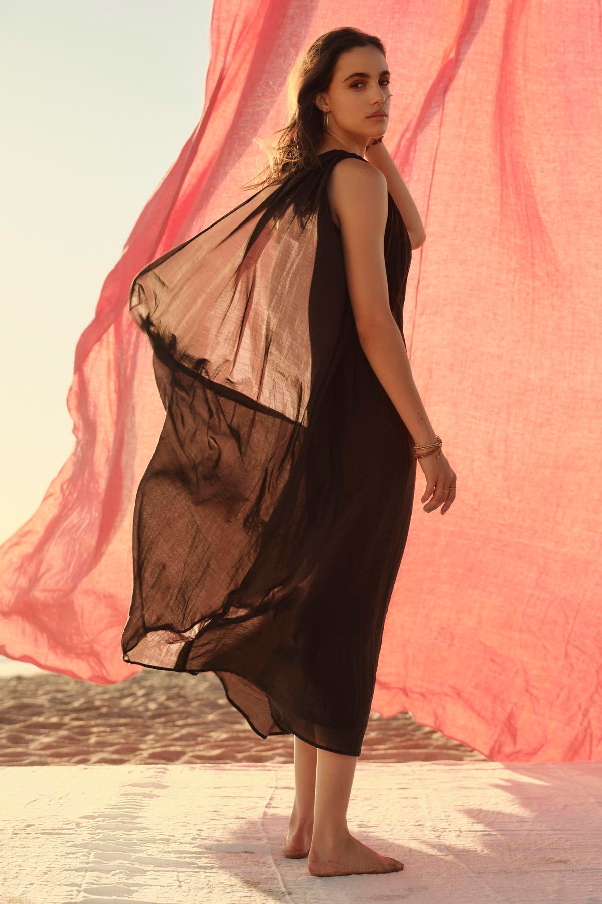 A woman in a flowing one-shoulder black Velvet by Graham & Spencer Diana dress stands barefoot, looking back, with a large pink fabric billowing in the background against a soft-lit sky.-36918819029185