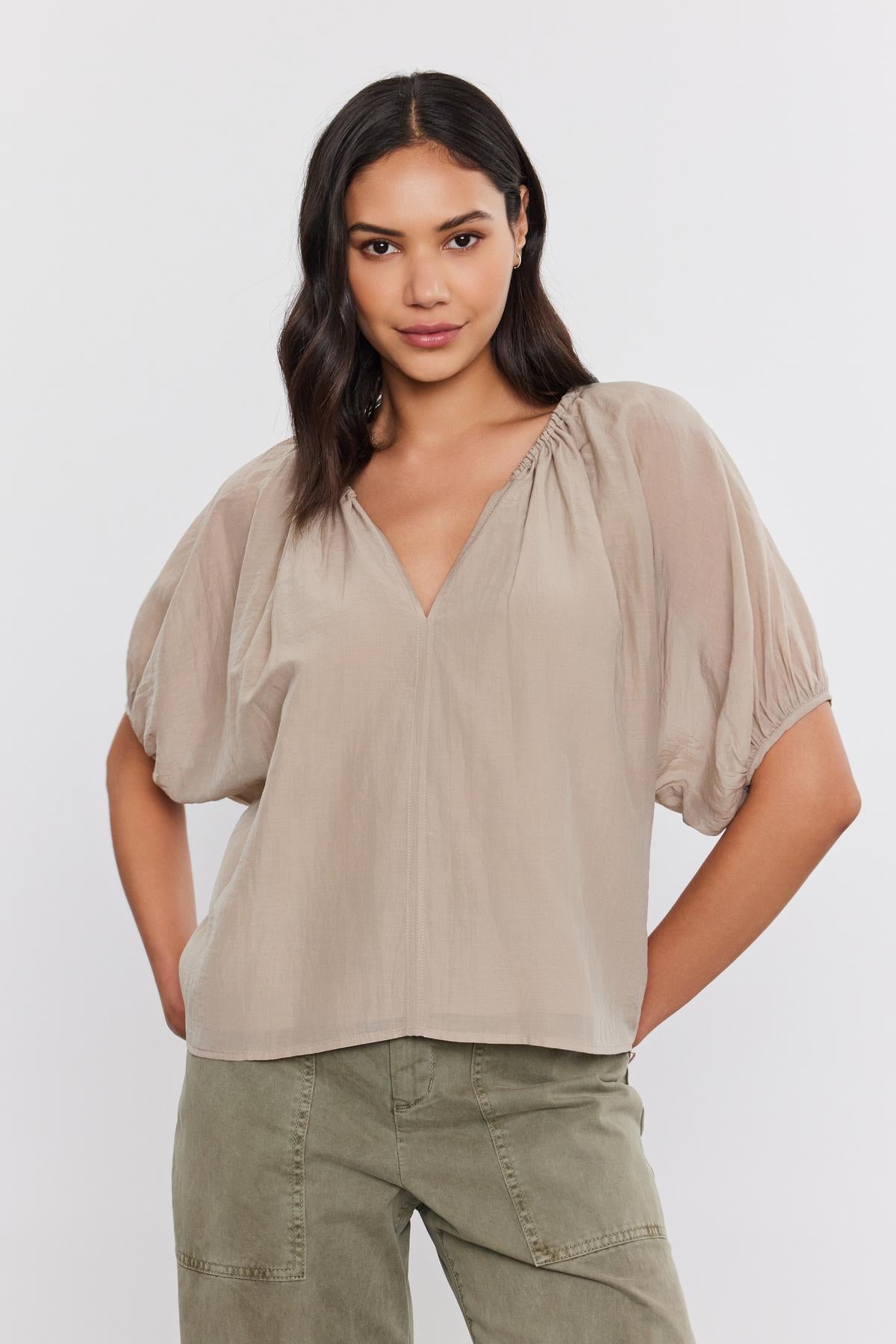   A person with shoulder-length dark hair is wearing the KRISTEN TOP in light brown and olive green pants from Velvet by Graham & Spencer. They stand with their hands on their hips against a plain white background. 