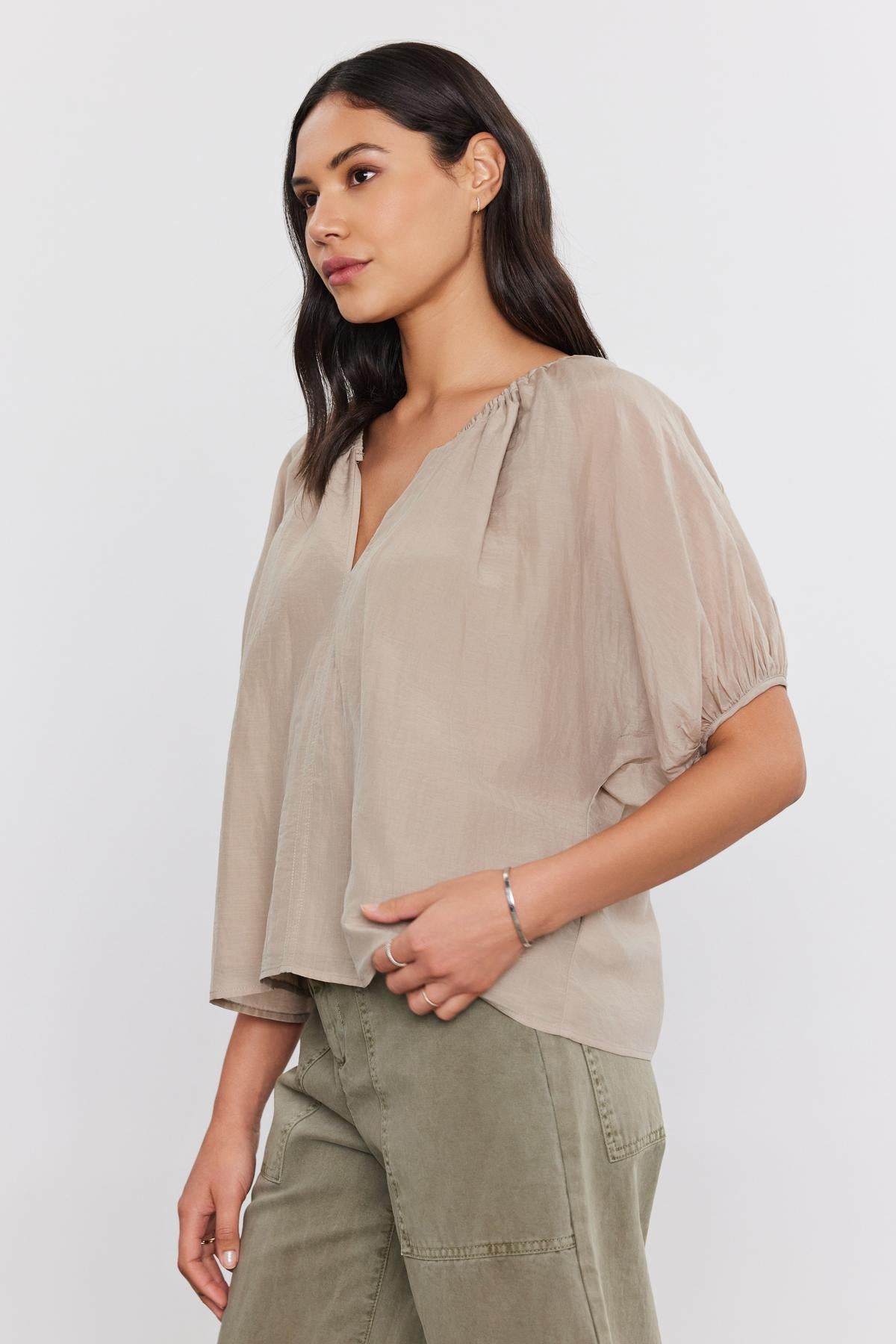   A woman with long dark hair, wearing the KRISTEN TOP by Velvet by Graham & Spencer—a light beige, loose-fitting blouse made of silk cotton voile with puff sleeves and a v-neckline—and olive green pants stands against a plain background, posing to the side. 