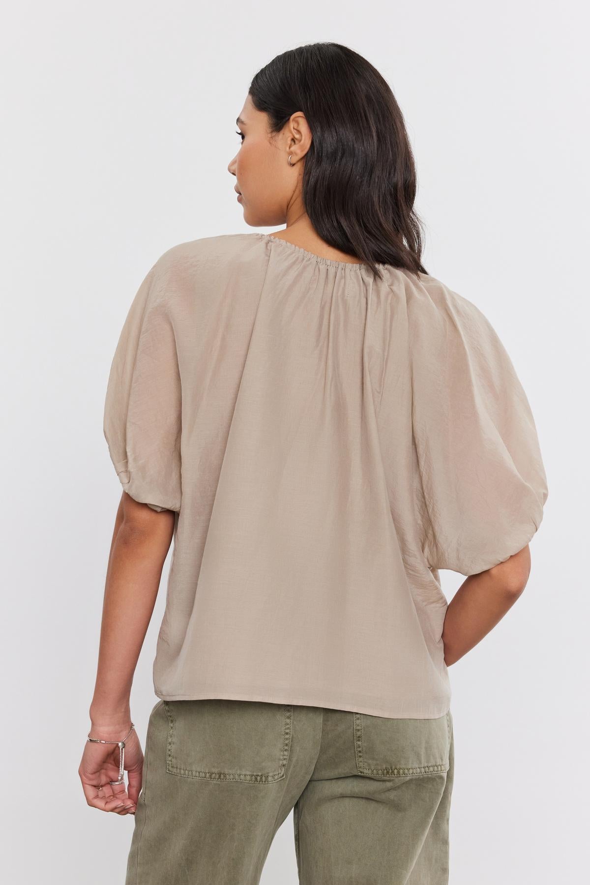   A woman with long dark hair wearing the KRISTEN TOP, a beige short-sleeve blouse with a v-neckline by Velvet by Graham & Spencer, and green pants is seen from the back. 