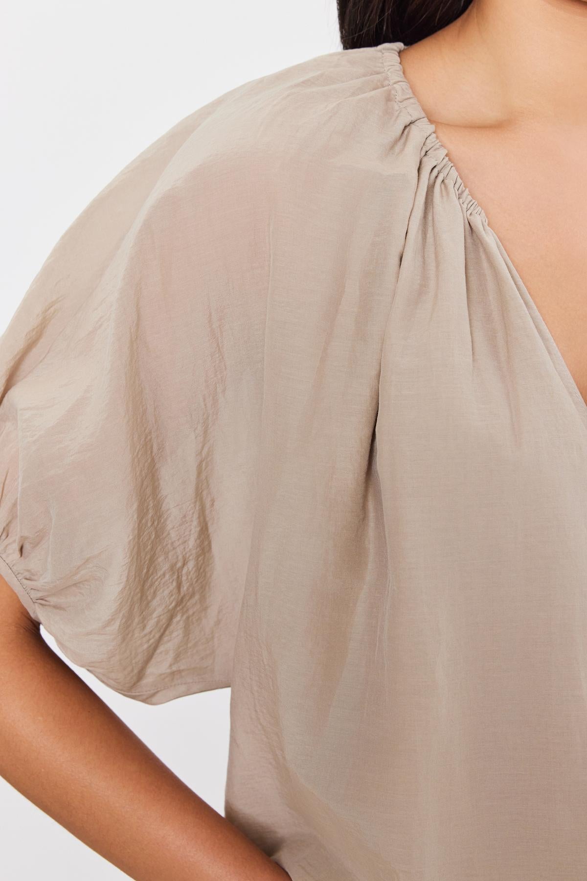   Close-up of a person wearing the Kristen Top from Velvet by Graham & Spencer. This light beige, loose-fitting blouse boasts gathered detailing near the shoulders and features delicate puff sleeves. Crafted from silk cotton voile, it also sports a subtle v-neckline. Only a portion of the blouse and part of the person's arm and hair are visible. 