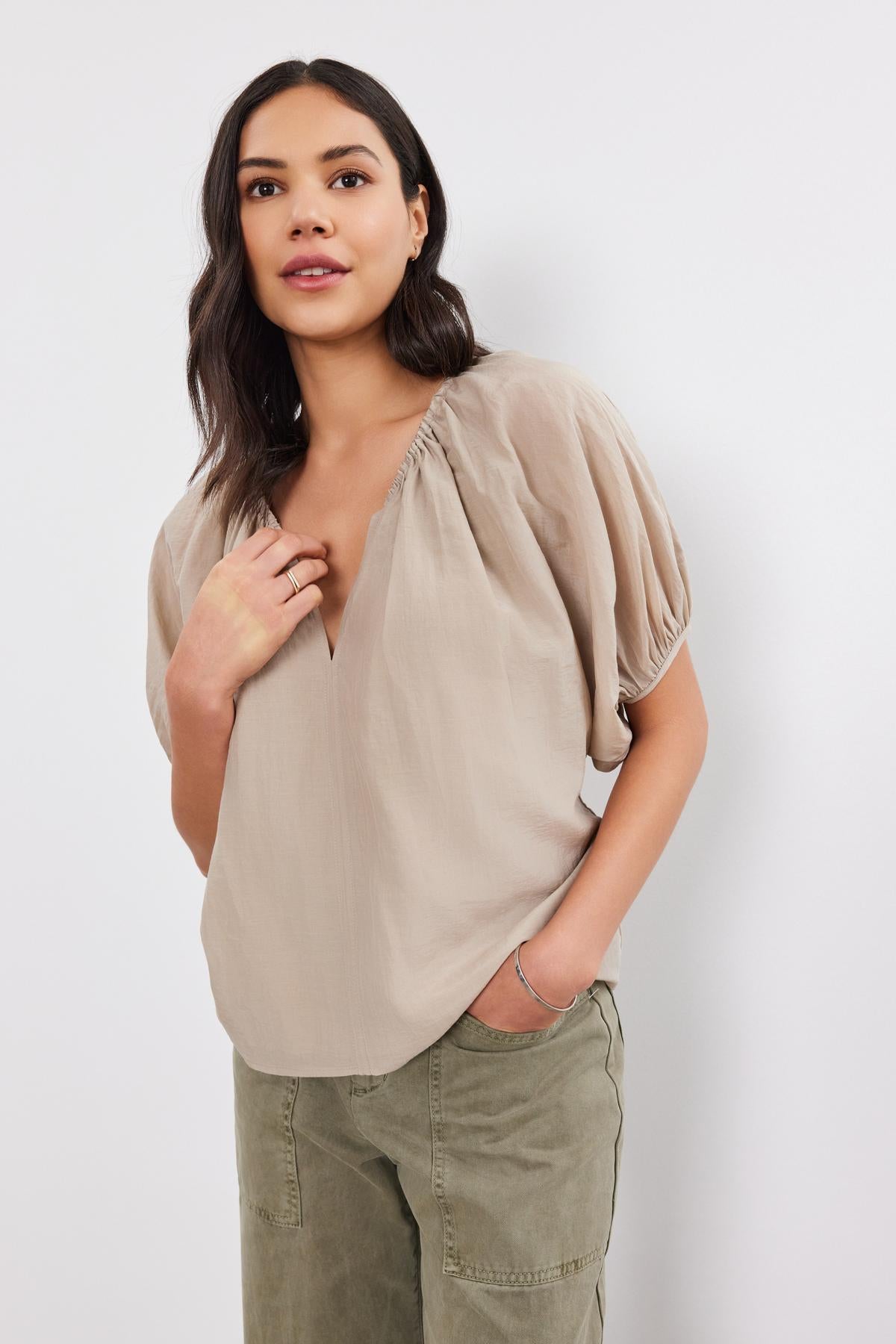   A woman stands against a white background, wearing the KRISTEN TOP by Velvet by Graham & Spencer, which features a taupe color with puff sleeves and a v-neckline. She has paired it with olive green pants, placing her right hand to her chest and her left hand in her pocket. 