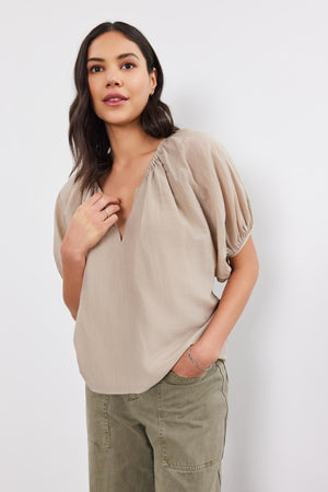 A woman stands against a white background, wearing the KRISTEN TOP by Velvet by Graham & Spencer, which features a taupe color with puff sleeves and a v-neckline. She has paired it with olive green pants, placing her right hand to her chest and her left hand in her pocket.