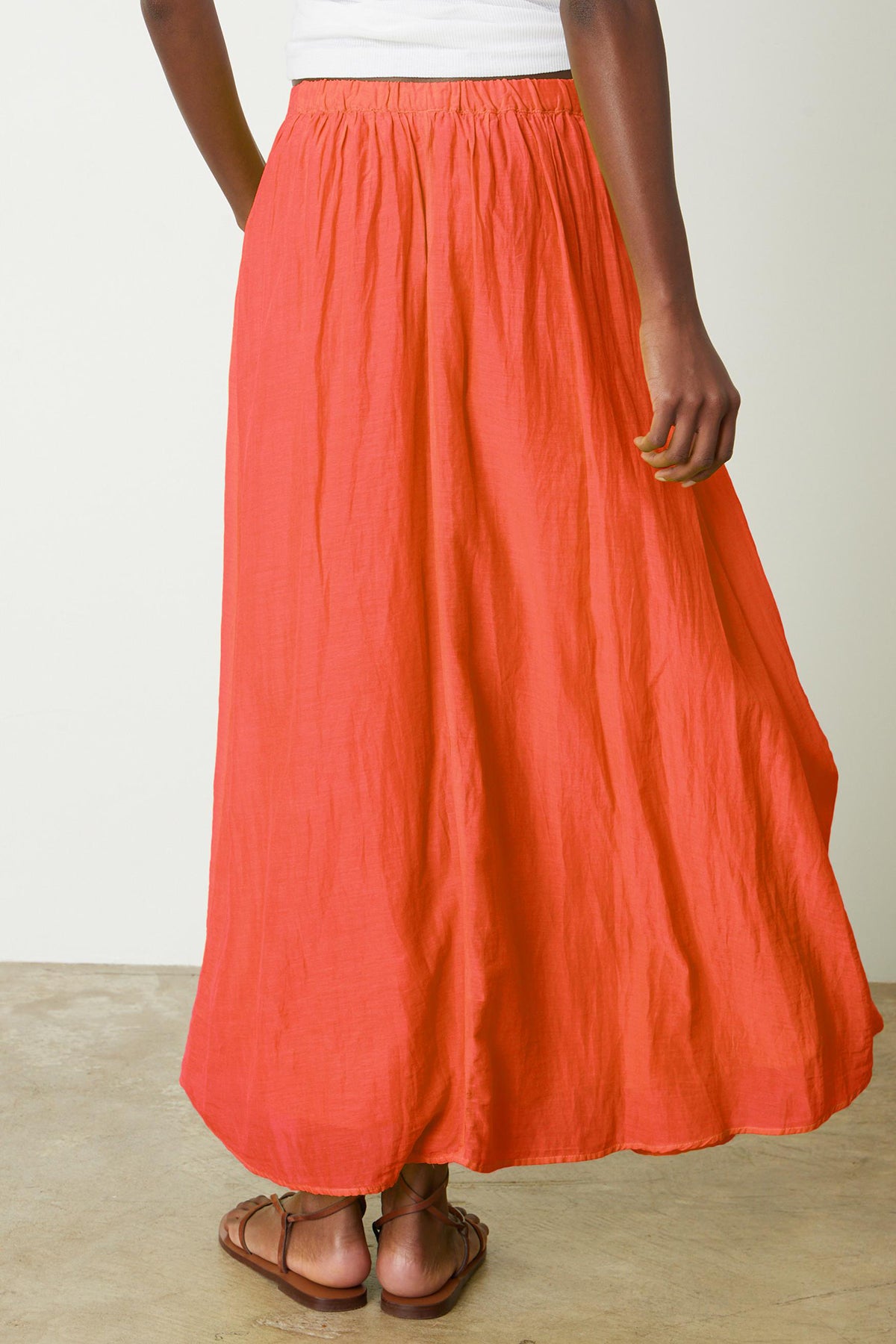   A person wearing a white top, the MARIELA SKIRT by Velvet by Graham & Spencer—an orange lined maxi skirt with pockets—and brown sandals stands on a concrete floor. The photo is taken from behind, showing the lower half of their body. 