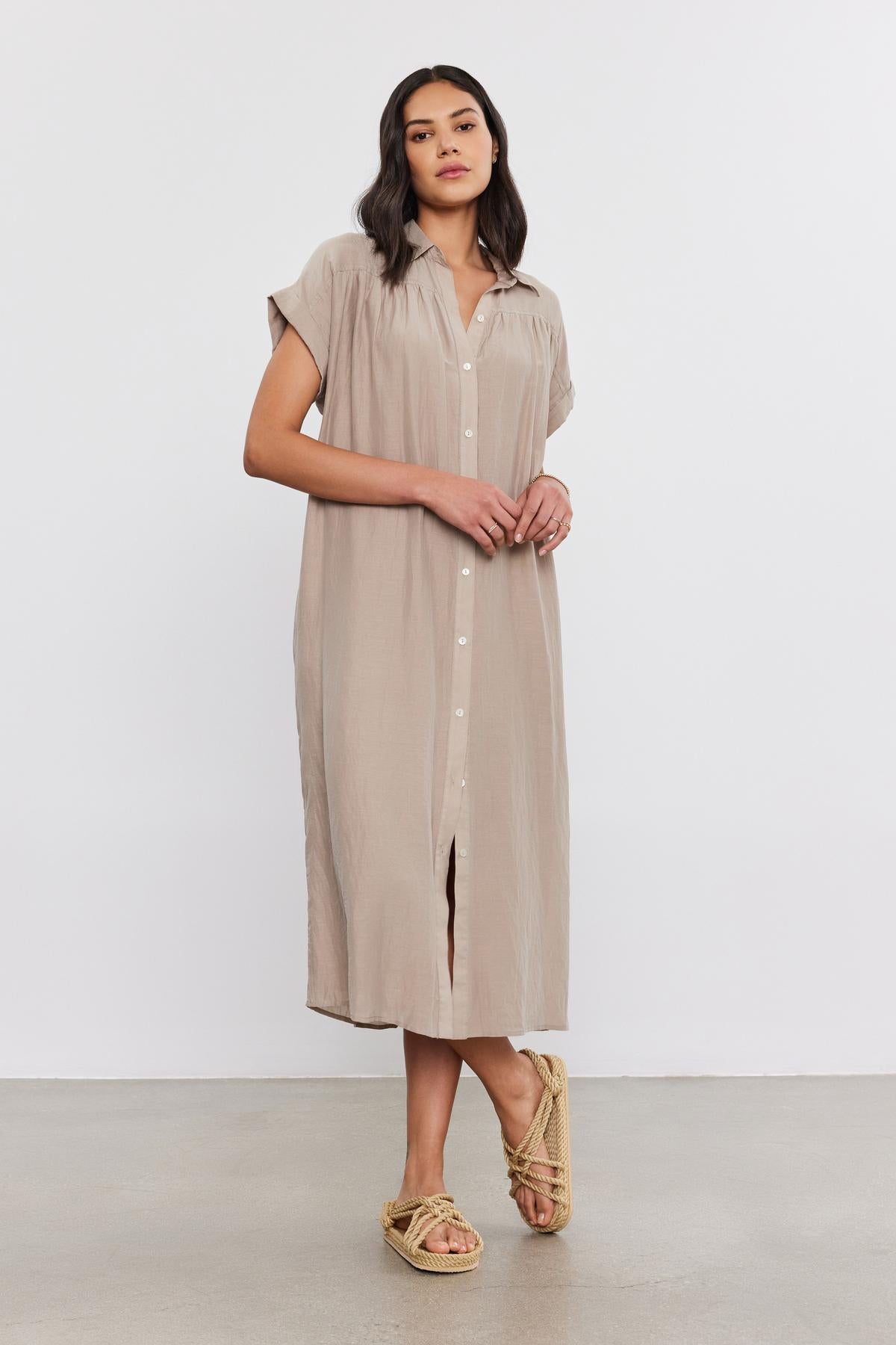   A woman stands against a plain background, wearing the MARLA SILK COTTON VOILE DRESS from Velvet by Graham & Spencer. The dress is loose-fitting and beige with short sleeves and buttons up the front. She completes her look with sandals and gazes directly at the camera, one hand resting on her hip. 