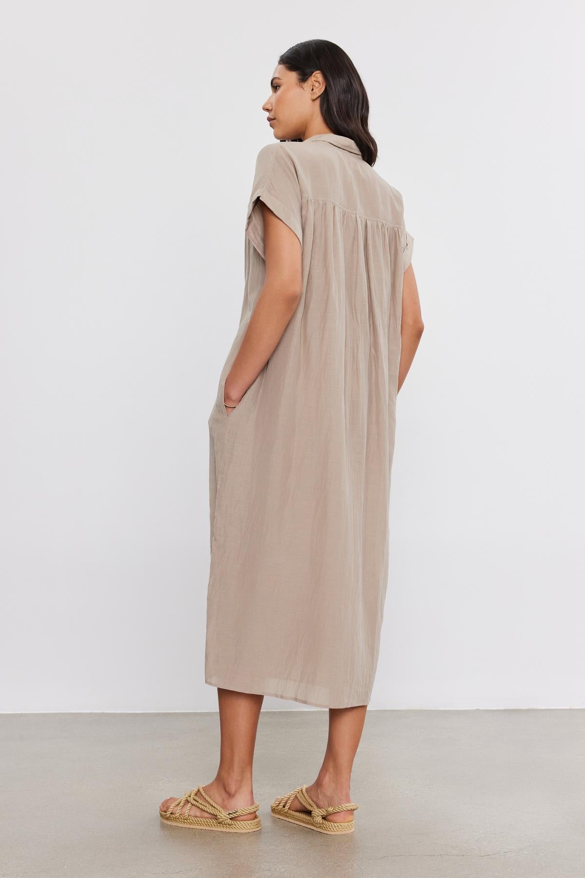   A person stands with their back turned, wearing the loose MARLA SILK COTTON VOILE DRESS by Velvet by Graham & Spencer and beige woven sandals. 