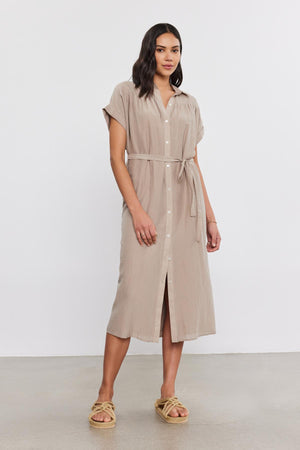 A woman stands against a plain white background, modeling the MARLA SILK COTTON VOILE DRESS by Velvet by Graham & Spencer. The beige, mid-length dress features short sleeves, button-down detailing, and a belt. She completes the look with tan braided sandals.