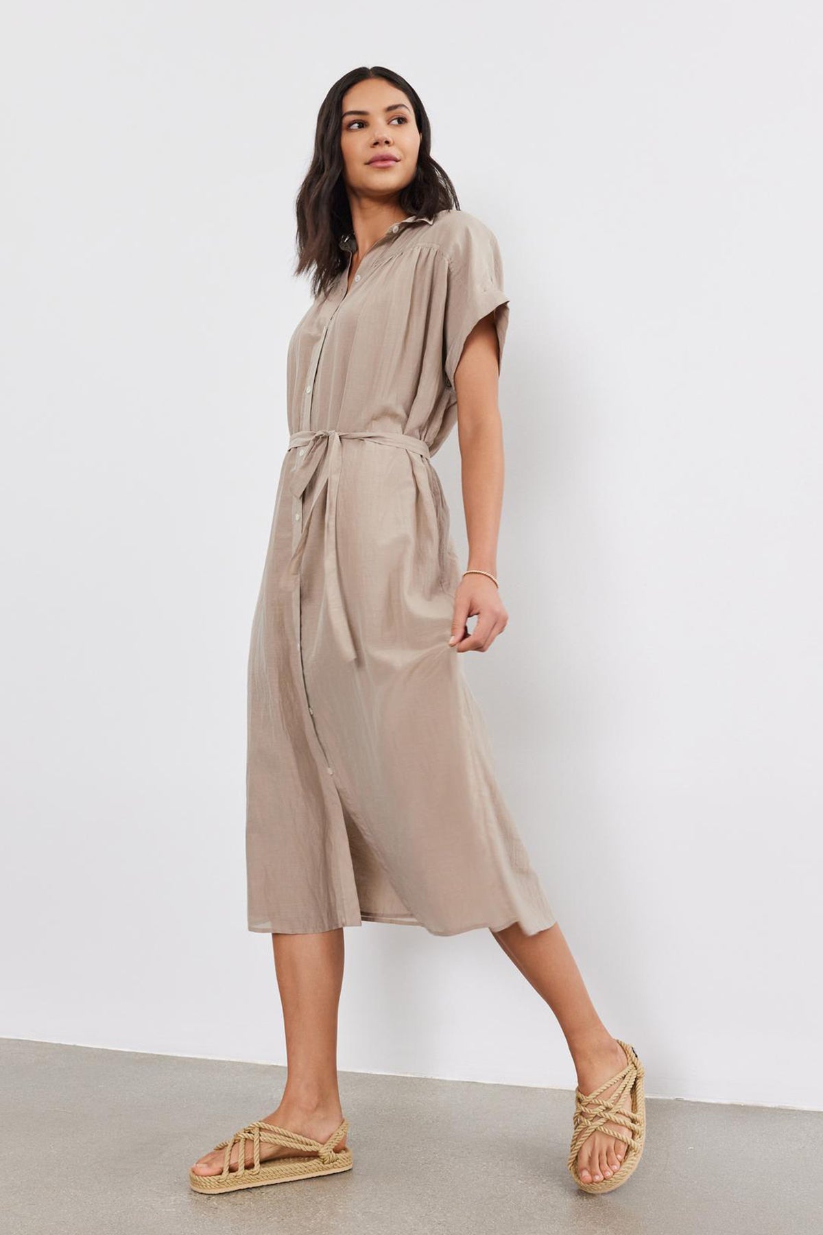   A woman in a beige MARLA SILK COTTON VOILE DRESS by Velvet by Graham & Spencer, featuring short sleeves, button-up detailing, and a waist tie, stands against a plain white background while wearing beige sandals. 