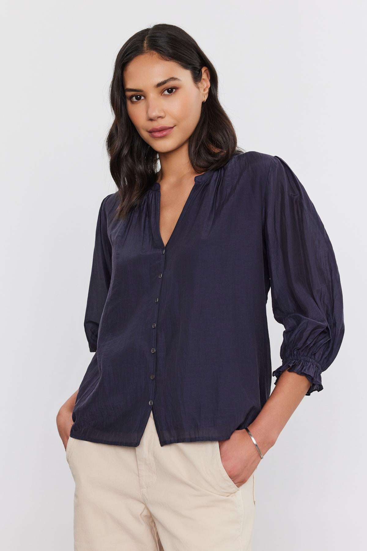   A woman with dark hair, wearing the MEL SILK COTTON VOILE BUTTON-UP TOP by Velvet by Graham & Spencer in navy and paired with light beige pants, is standing against a plain white background. 