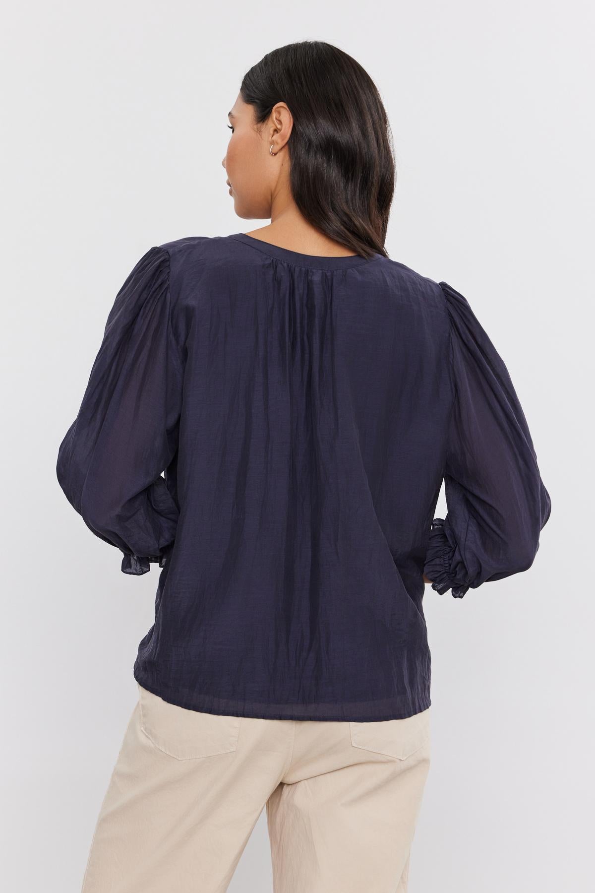   A person is modeling the Velvet by Graham & Spencer MEL Silk Cotton Voile Button-Up Top with long puff sleeves and light-colored pants, standing with their back to the camera. 