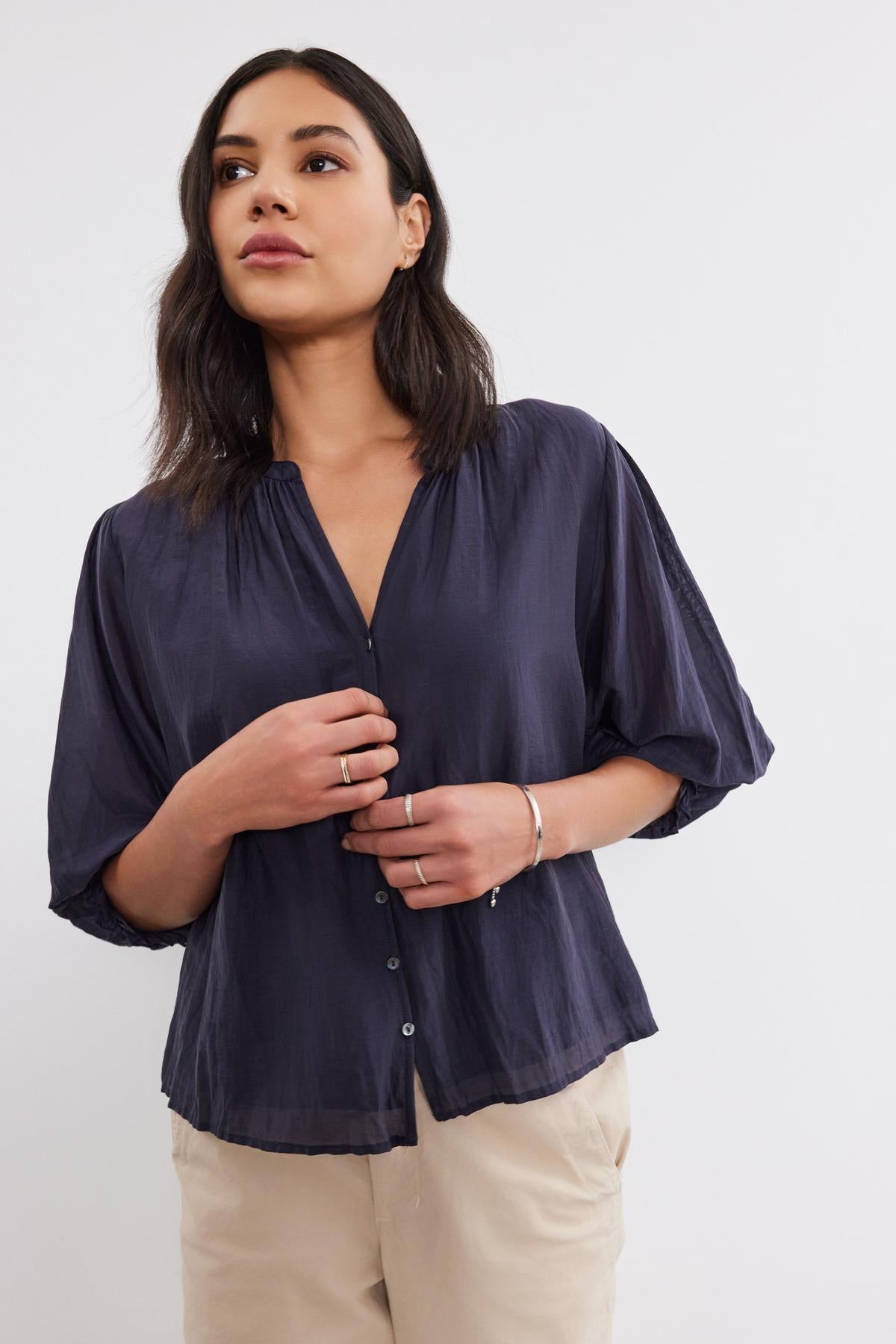   A person with long, dark hair stands wearing a MEL SILK COTTON VOILE BUTTON-UP TOP by Velvet by Graham & Spencer and beige pants. They have rings and a bracelet on their left hand. 