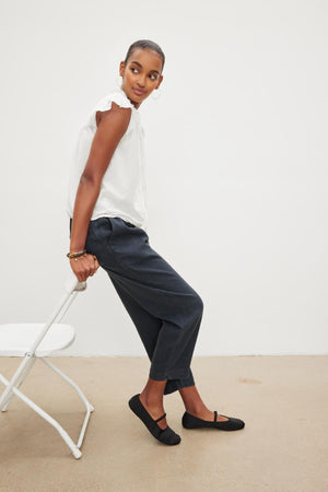 A woman is leaning against a chair in a MELANIE V-NECK BLOUSE by Velvet by Graham & Spencer in white shirt and black pants.