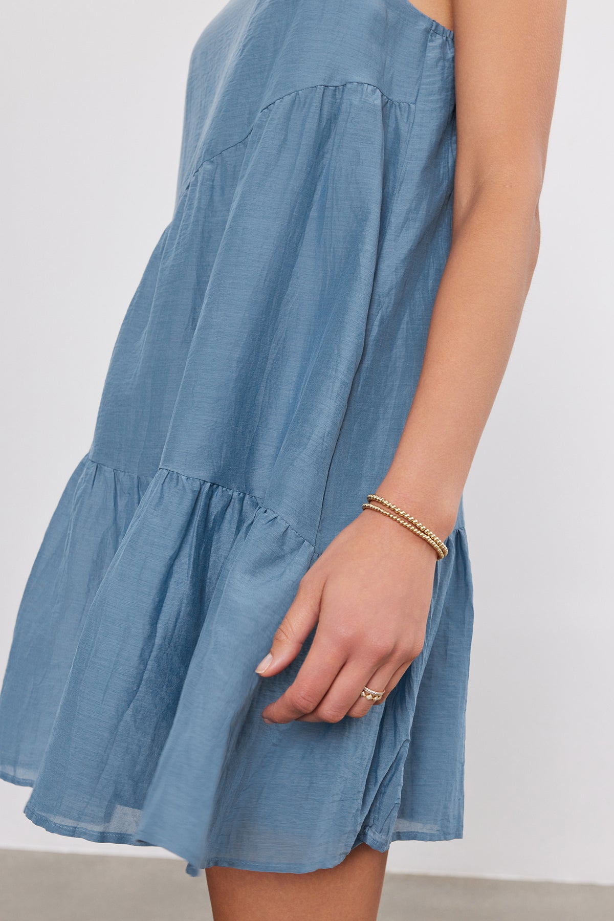   A person wearing a blue asymmetrical tiered SUZIE DRESS by Velvet by Graham & Spencer and a gold bracelet, showing their arm and some of the dress's fabric. 
