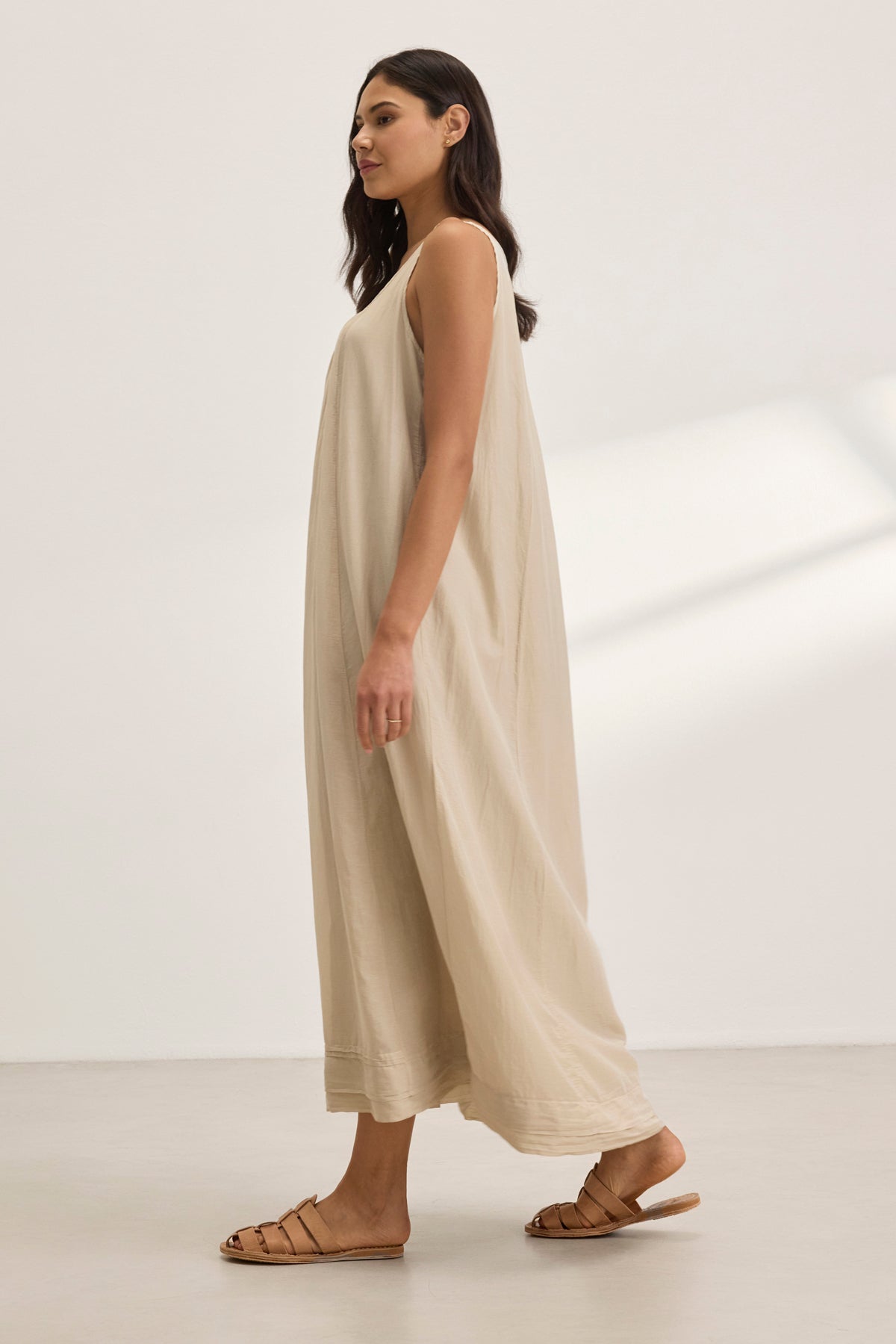 A woman in a BAYOU SILK COTTON VOILE DRESS by Velvet by Graham & Spencer, featuring a scoop neckline, stands against a plain, light background. The elegant beige maxi dress gracefully drapes down to her ankles, paired with brown sandals.-38801131274433