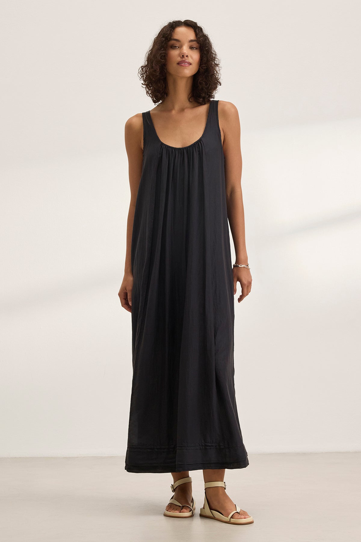   A woman wears the BAYOU SILK COTTON VOILE DRESS by Velvet by Graham & Spencer—a sleeveless, loose-fit maxi with a detachable belt—paired with light sandals, standing indoors against a plain background. 