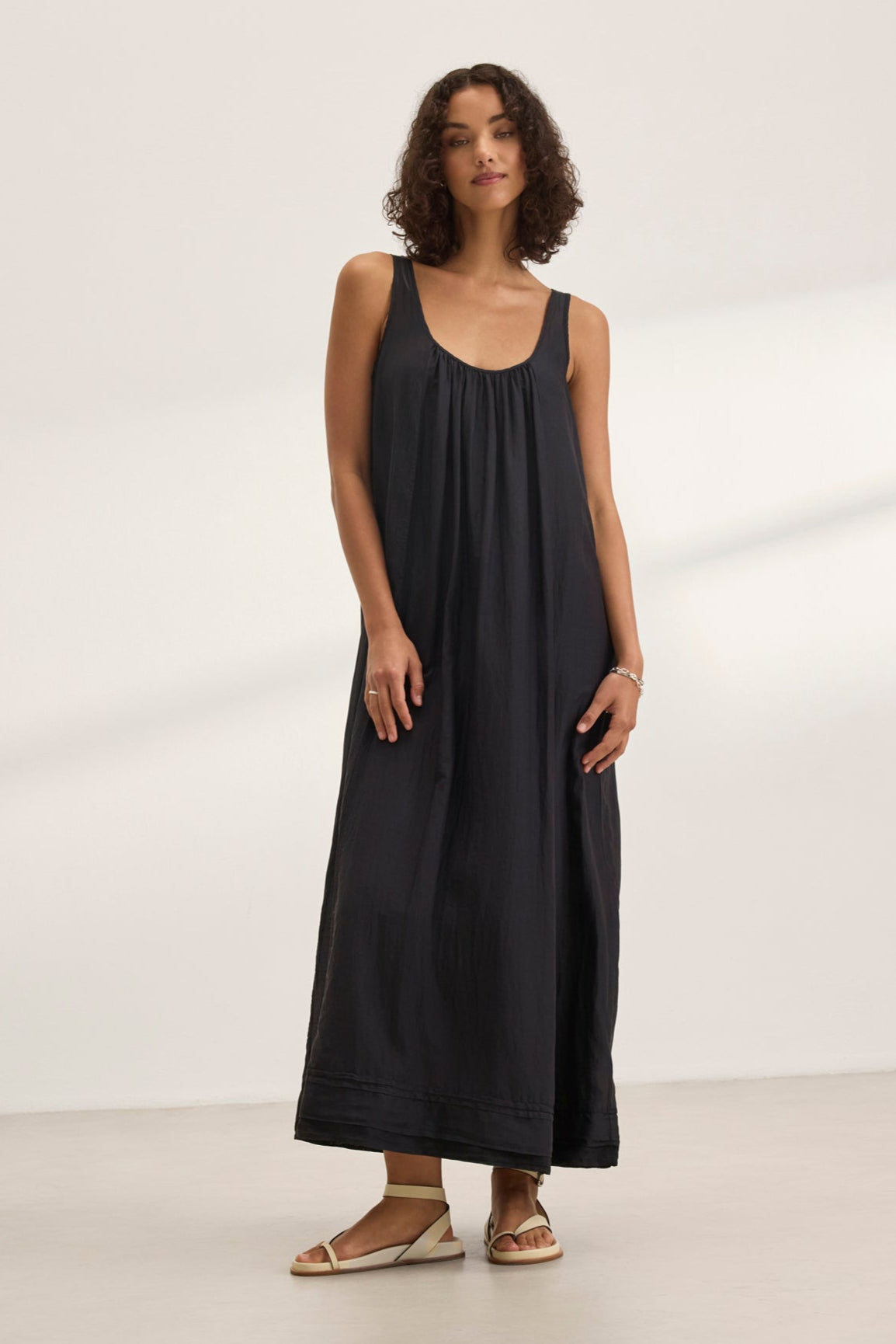 A woman showcases the BAYOU SILK COTTON VOILE DRESS by Velvet by Graham & Spencer, a sleek sleeveless black maxi, paired with sandals. She stands on a light-colored floor against a white background, highlighting its versatile styling perfect for any occasion.-38801131012289